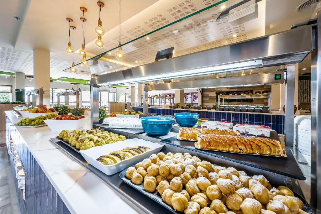 Bagātīgs brokastu bufete ar dažādiem augļiem, konditorejas izstrādājumiem un desertiem, kas izvietoti uz elegantas marmora virsmas. Gaisīga atmosfēra ar modernu interjeru un stilīgām lampām.