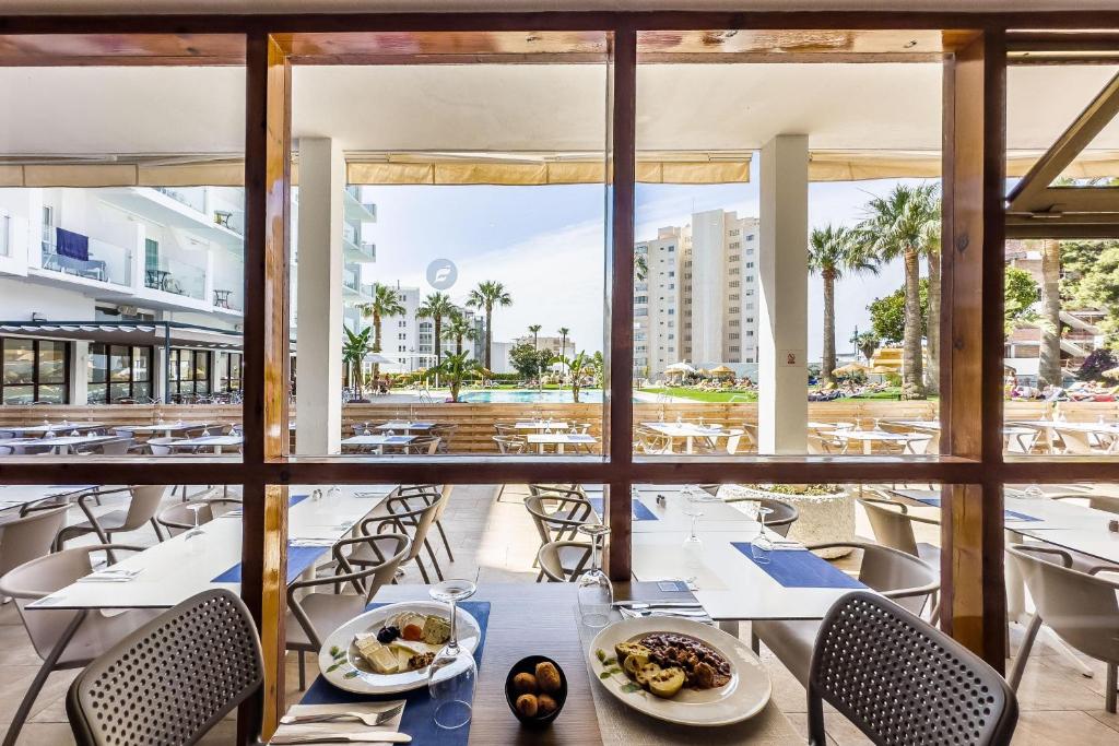 Restorāna skats ar ēdienu uz galda, kas vērsts uz peldbaseinu un apkārtējiem palmām. Gaisīga atmosfēra un moderns interjers.