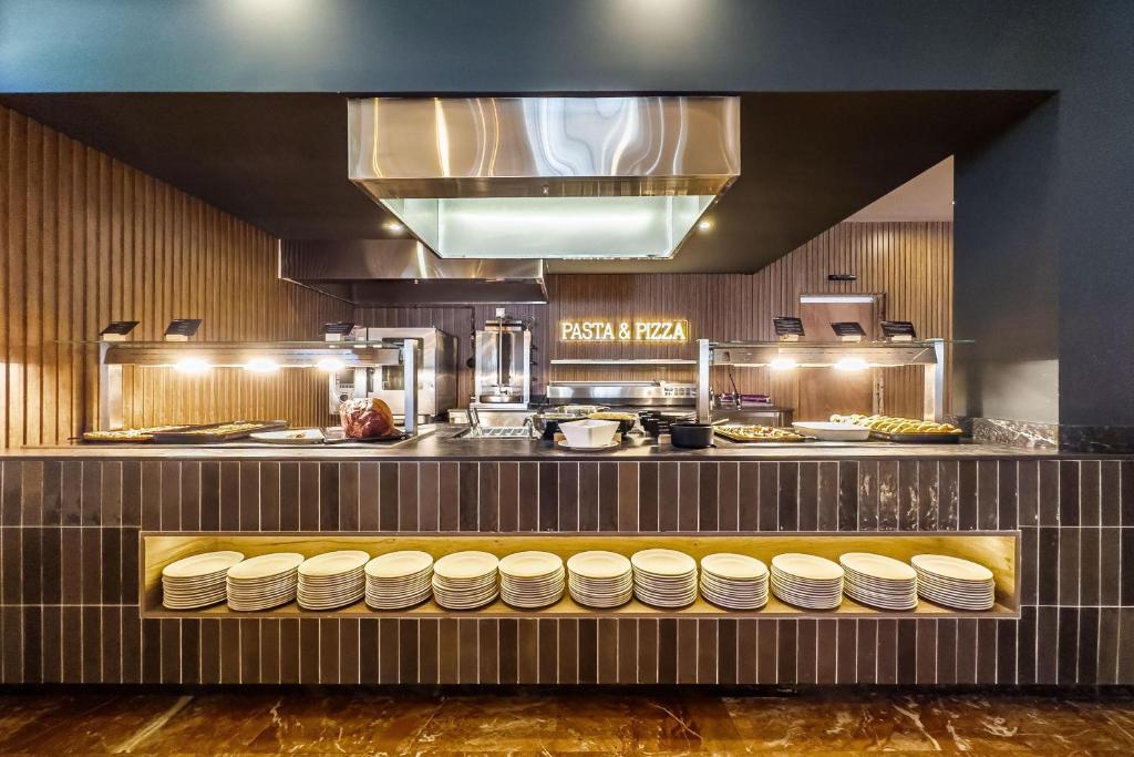 Mūsdienīgs restorāna bārs ar "Pasta & Pizza" zīmi, kur redzamas dažādas ēdienu uzkodas un sakrautas šķīvju rindas.