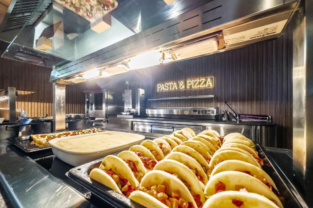 Restorānā redzami dažādi ēdieni, tostarp tacos un pica, ar izgaismoto uzrakstu "PASTA & PIZZA" fonā.