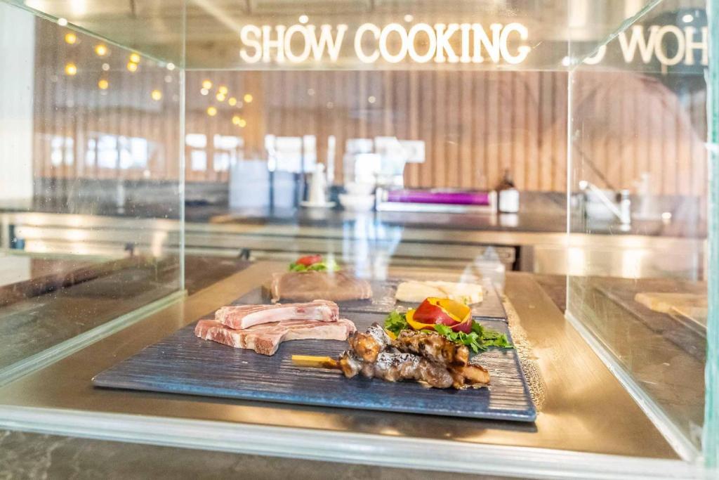 Šajā attēlā redzama izsmalcināta gaļas plāksne, kas izkārtota uz melnas virsmas, ar dārzeņiem un augļiem. Fons ietver modernu virtuvi ar gaismu un "Show Cooking" uzrakstu.
