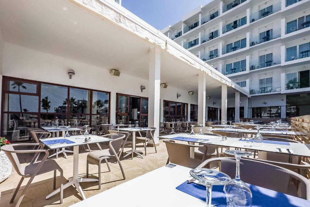 Mūsdienīgs restorāns ar terasi, kurā ir galdi un krēsli, gaida viesus. Apskatāma viesnīcas fasāde un saulaina atmosfēra.