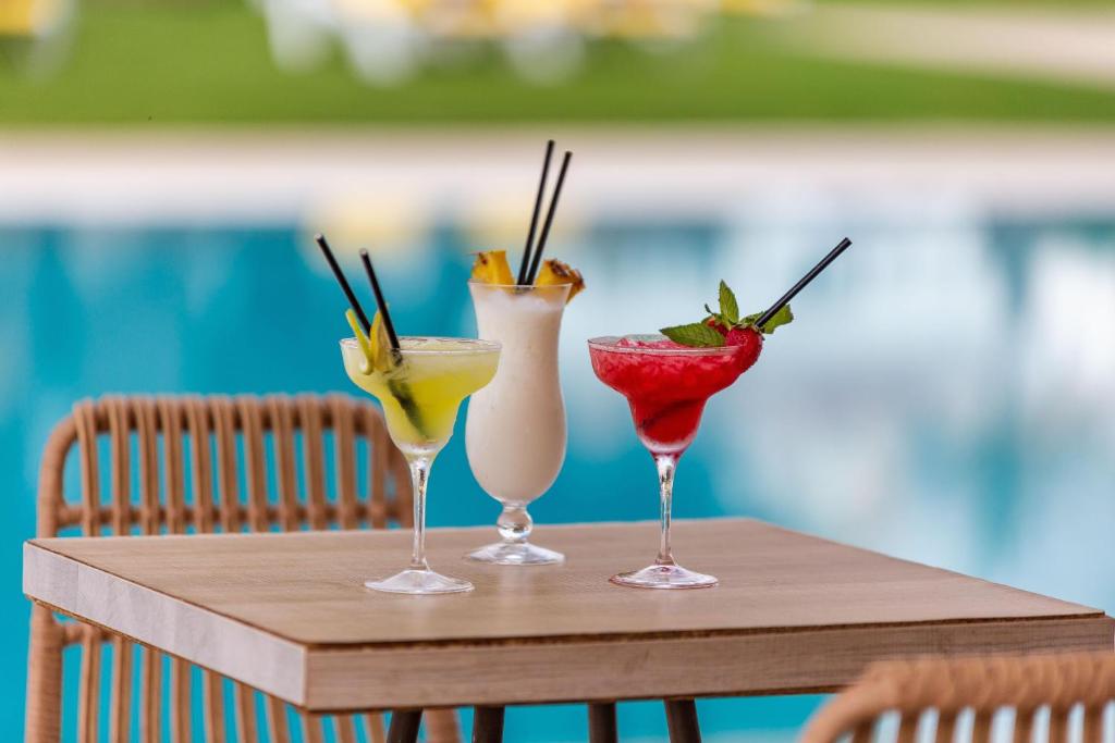 Trīs krāsaini kokteiļi uz koka galda pie baseina, radot vasarīgu un relaksējošu atmosfēru.
