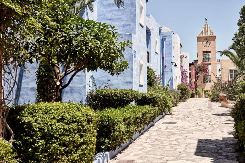 Skaists, šaurais ceļš ar ziliem un krēmkrāsas māju fasādēm, ko ieskauj zaļi krūmi un palmas, rada mierīgu atmosfēru. Redzama arī pulksteņa torņa struktūra, kas piešķir ainavai raksturu.