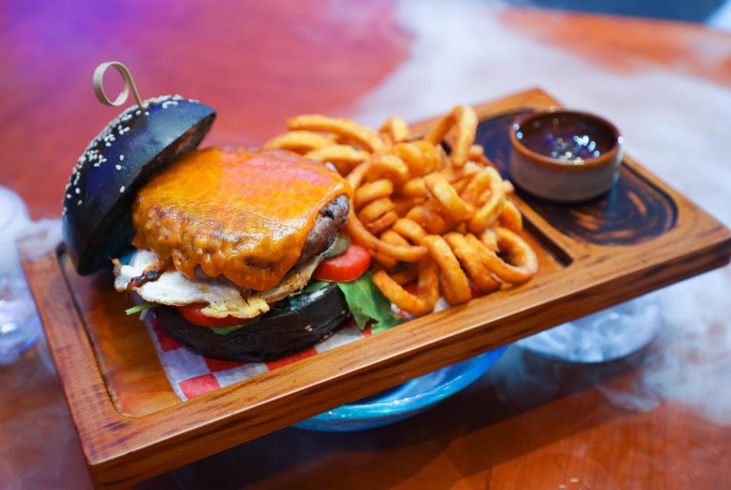 Burger ar melno maizi, siera kārtu un ceptiem kartupeļiem, pasniegts uz koka šķīvja ar mērci.