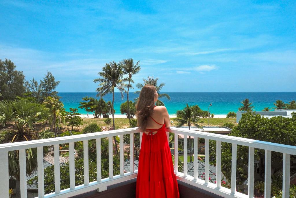 Sieviete sarkanā kleitā stāv uz balkona, skatoties uz skaisto pludmali un zilo jūru, ko ieskauj palmas. Ideāls brīvdienu brīdis tropiskā paradīzē.