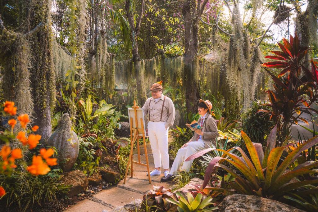 Dārzs ar zaļām augu kompozīcijām, kur mākslinieks zīmē uz eļļas audekla, bet otra persona lasa grāmatu. Apkārt krāšņas puķes un dabiska vide.