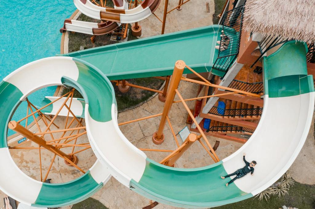 Ūdens slidkalniņš ar zaļām un baltām līnijām, kas ved uz peldbaseinu, ar cilvēku, kas gatavojas nobraukt. Apkārt redzams tropisks ainavas fons.