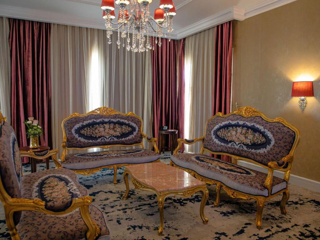 Elegants viesistabas interjers ar zelta apdari, greznu mēbeļu komplektu un krāšņām aizkariem. Centrā atrodas stilīgs kafijas galdiņš, radot patīkamu atmosfēru.