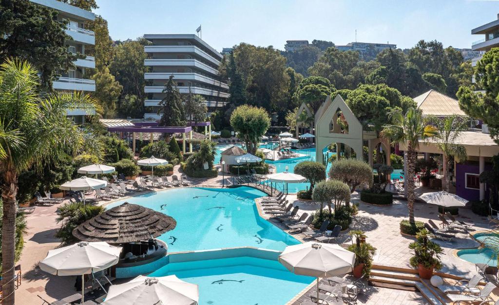 Lieliska kūrorta baseina zona ar palmām un sauļošanās krēsliem, kas izvietoti apkārt. Redzams moderns viesnīcu komplekss ar zaļu dabu un relaksējošu atmosfēru.