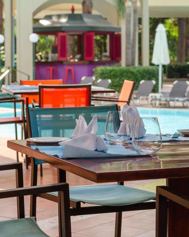 Restorāna galds ar eleganti salocītām salvetēm un glītām glāzēm, fona ainavā redzams peldbaseins un krāsains bārs.
