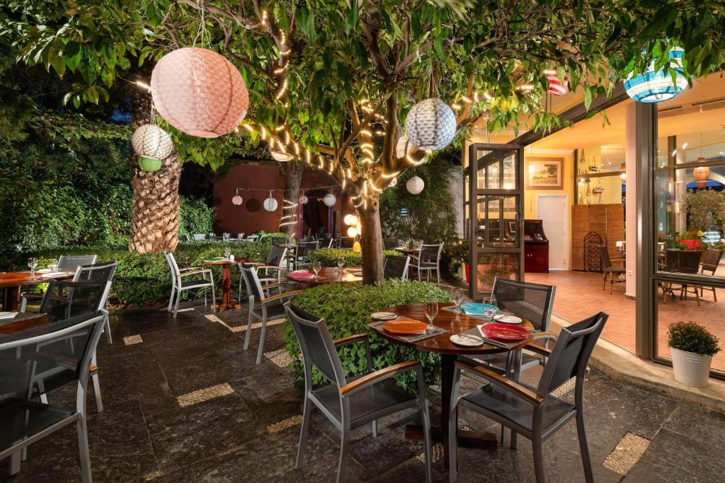 Āra restorāna terase ar apgaismotām laternām un galdiem, kas izkārtoti zem zaļojoša koka. Atpūtas zona ar augu rotājumiem un mājīgu atmosfēru.