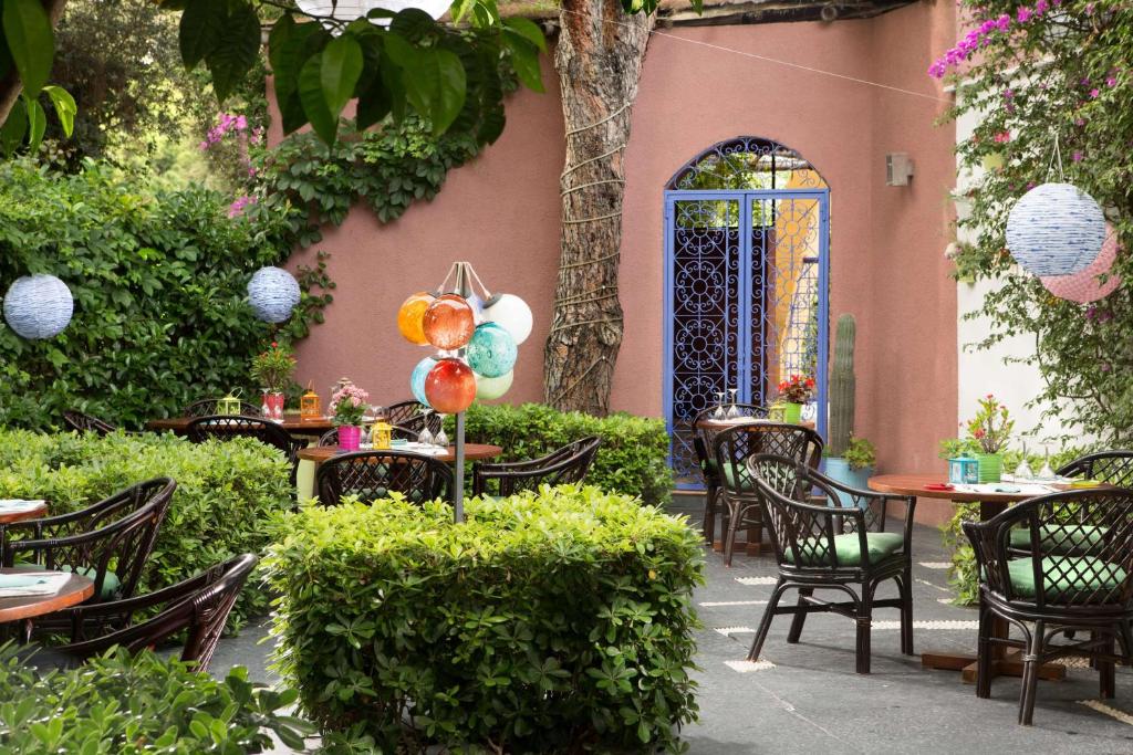 Kāds zaļš dārzs ar koka galdiem un krēsliem, kur piekārti krāsaini laternas un ziedi, radot patīkamu atmosfēru. Aiz krūmiem redzams zils durvis ar dekoratīvu režģi.