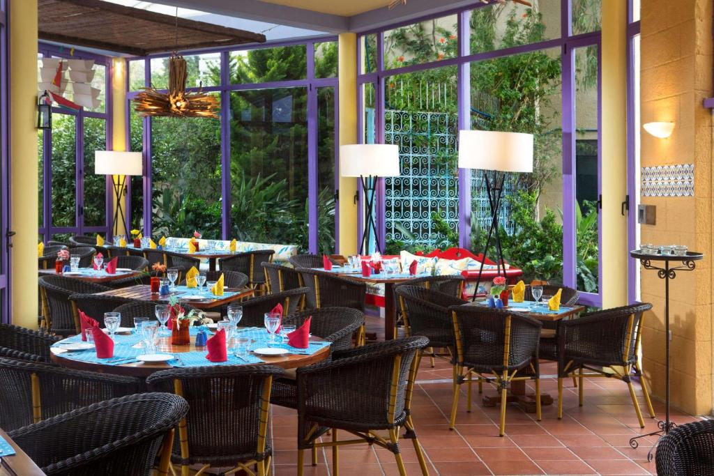 Mūsdienīgs restorāns ar plašām logu sienām, kas nodrošina dabiska apgaismojuma plūsmu. Galdi ir klāti ar krāsainiem salvetēm un galda piederumiem, radot dzīvīgu un viesmīlīgu atmosfēru.