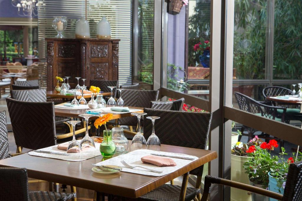 Elegants restorāna interjers ar koka galdiem un krēsliem, izrotāts ar ziediem un glāzēm, kas rada patīkamu atmosfēru. Labi apgaismots, ar zaļumiem un ziediem ārpus logiem.