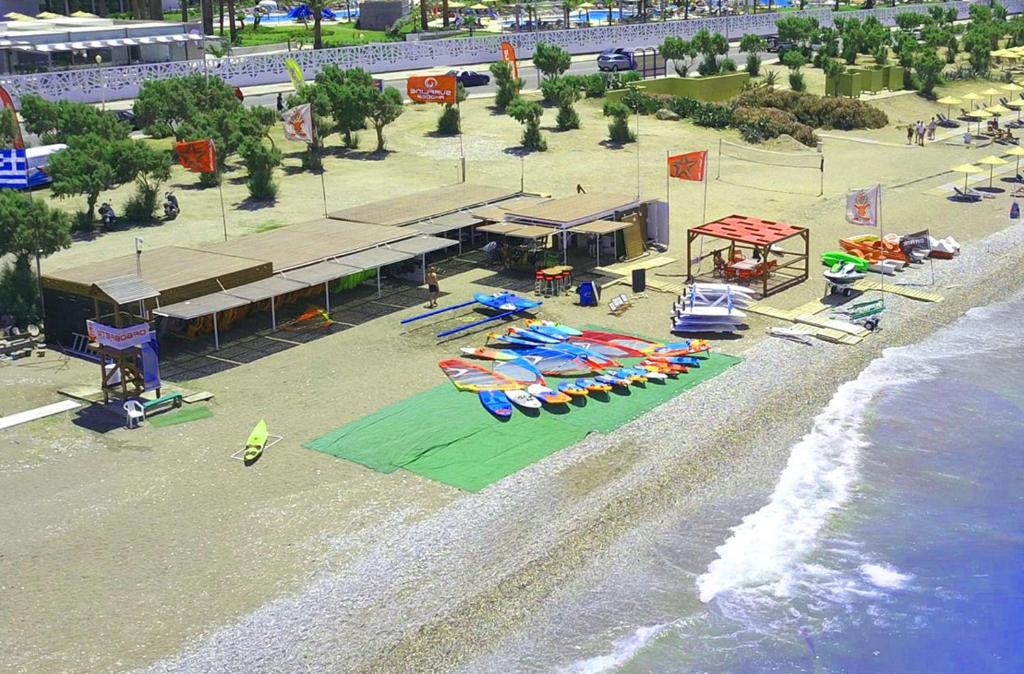 Pludmales aktivitāšu centrs ar krāsainām laivām un ūdens sporta inventāru, kas izvietots smilšu pludmalē. Apkārt redzami koki un pludmales struktūras.