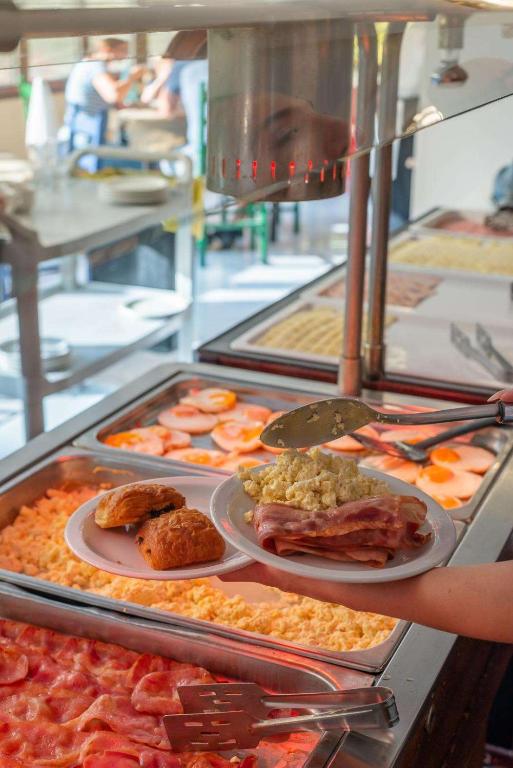 Brokastu bufete ar dažādām ēdienu izvēlēm, tostarp olām, šķiņķi un kruasānu. Fonā redzami apmeklētāji, kas bauda maltīti.