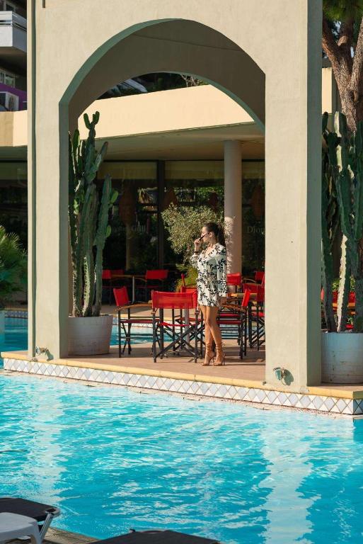 Sieviete stāv zem arkas pie peldbaseina, tērpta vieglā apģērbā un tur rokās dzērienu. Fons ietver kaktusus un sarkanas krēslus.