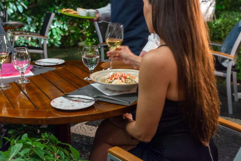 Eleganta vakariņu aina ar cilvēkiem pie galda, kur pasniegts salāti un vīns. Apkārt ir zaļa vide, radot patīkamu atmosfēru.