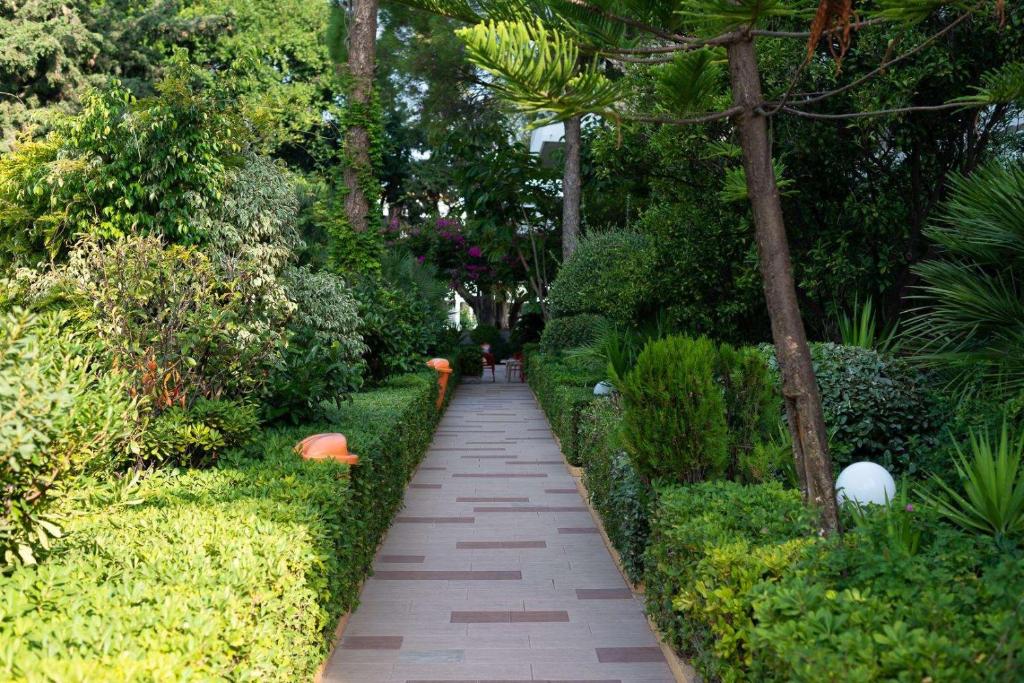 Zaļais dārzs ar bruģētu celiņu, apkārt augošu krūmu un koku daudzveidību, kas rada mierīgu un relaksējošu atmosfēru. Celiņš ved uz noslēpumainu vietu, ko ieskauj ziedi un apgaismojuma elementi.