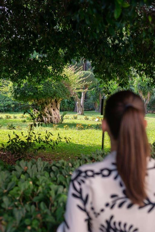 Sieviete ar matiem ponijā, tērpta rakstainā apģērbā, vēro dārza ainavu ar zaļu zālienu un kokiem. Daba ap viņu ir zaļa un dzīvīga, radot mierīgu atmosfēru.