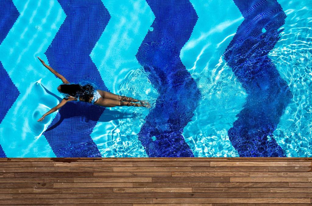 Peldētāja, kas brīvi kustas pa zilo baseinu ar viļņainu rakstu, radot burbuļus un atspīdumus ūdenī. Baseina apmalē redzama koka dēļu virsma.