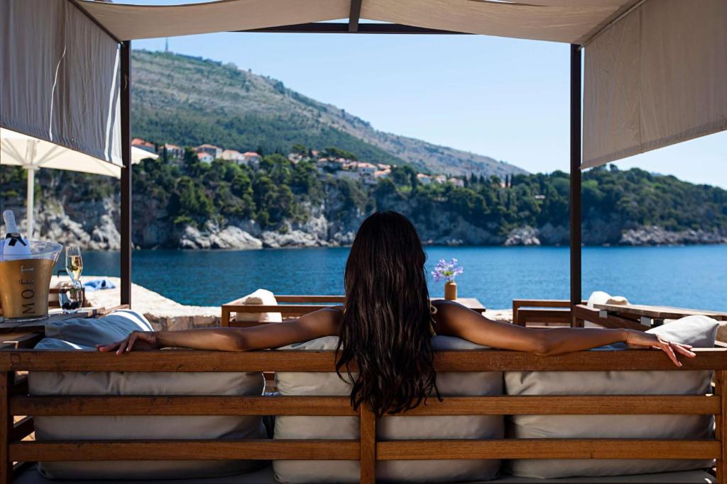 Relaksējoša ainava pie jūras, kur sieviete sēž uz dīvāna un bauda skatu uz kalniem un ūdeni. Uz galda atrodas šampanieša pudele un glāze.