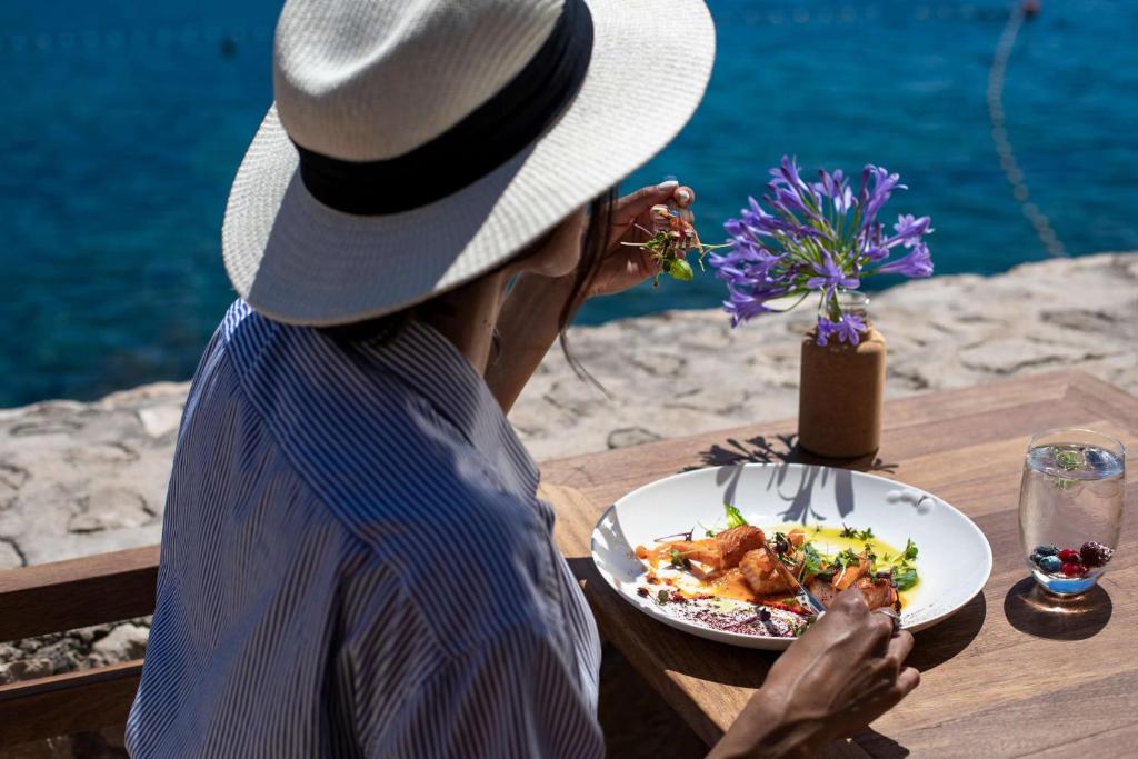 Sieviete ar platmali sēž pie galda, baudot ēdienu pie jūras ar ziediem vāzē. Svaiga maltīte un dzēriens papildina relaksējošo atmosfēru.