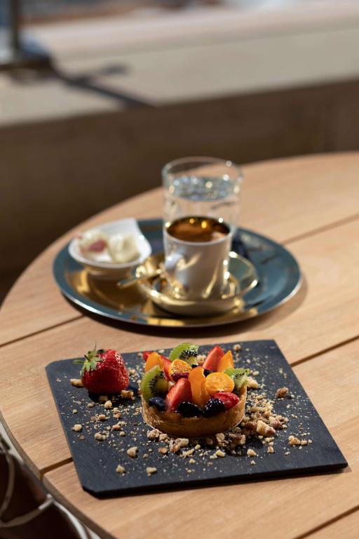 Svaiga augļu torte uz melna šķīvja, blakus tase kafijas un ūdens glāze uz koka galda.