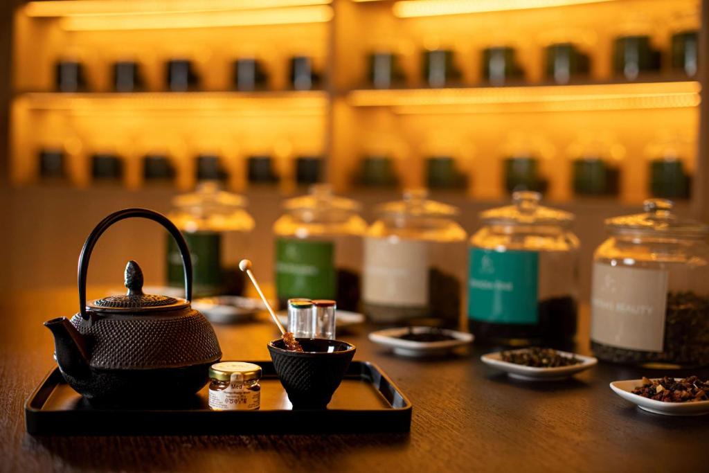 Melnā tējas kanna atrodas uz galda, blakus sīkiem traukiem ar dažādām tēju šķirnēm un garšvielām. Fons ir mirdzošs ar izkārtotām burkām, kas satur dažādas tējas.