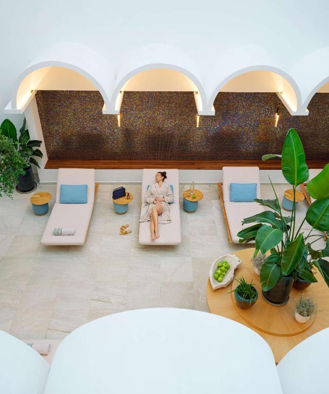 Mūsdienīgs spa telpas skats ar guļamkrēslām, kurās atpūšas sieviete, kā arī augiem un dekoratīviem elementiem. Telpā ir harmoniska atmosfēra, kas veicina relaksāciju.