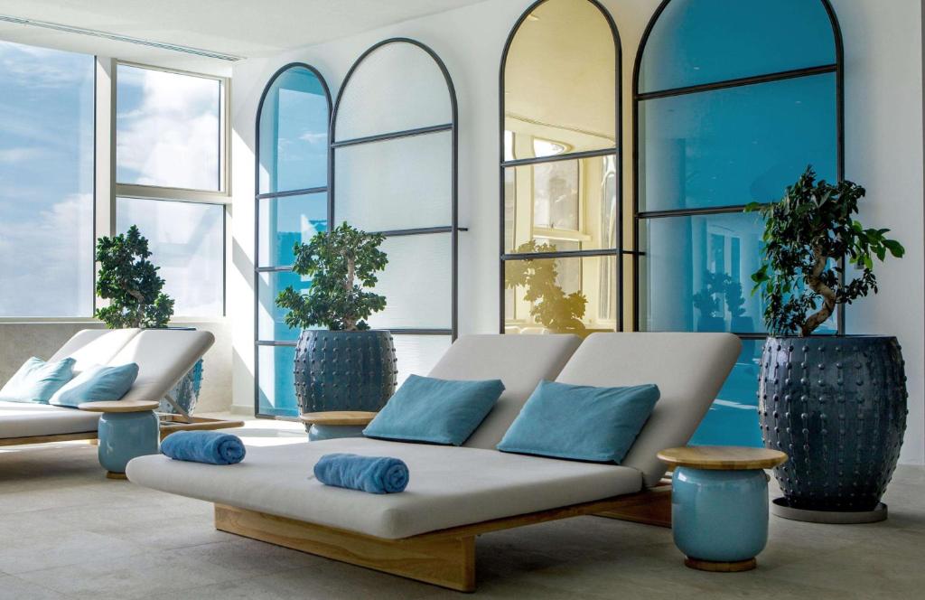 Mūsdienīga spa telpa ar ērtām guļamvietām, zilām akcentiem un zaļām augiem, kas rada relaksējošu atmosfēru. Lielie logi ļauj dabīgajai gaismai ieplūst telpā.