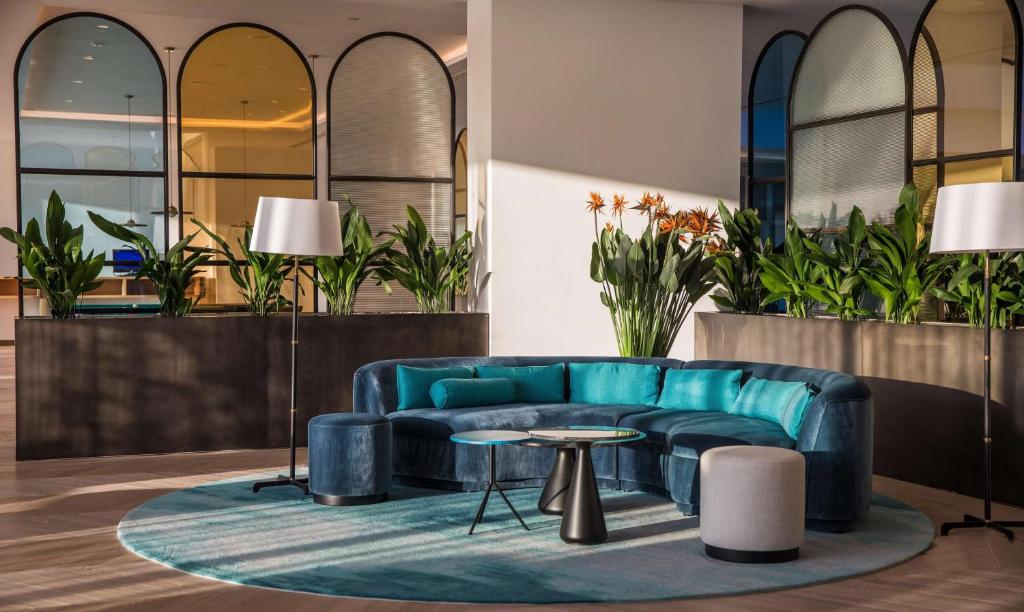 Mūsdienīga viesistaba ar stilīgu zilo dīvānu, apaļu paklāju un augiem, kas rada svaigu atmosfēru. Gaismas avoti un lieli logi piešķir telpai dzīvīgumu.