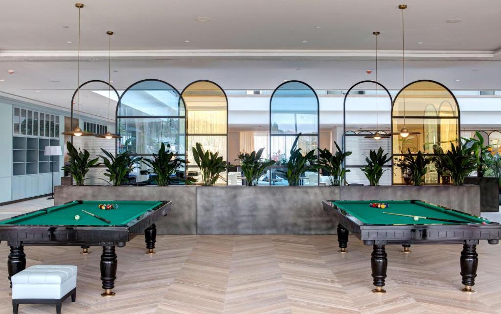 Mūsdienīga biljarda zona ar zaļām biljarda galdiņām un augstiem logiem, kas ļauj ienākt dabiskajai gaismai. Interjers papildināts ar zaļām augiem un elegantu apgaismojumu.