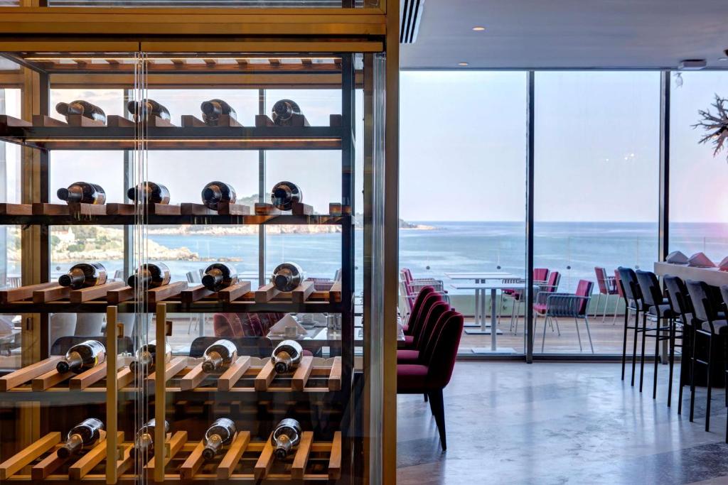 Eleganta restorāna interjers ar vīna plauktu un skatu uz jūru. Vīna pudeles ir sakārtotas uz koka plauktiem, bet fonā redzama atvērta terase ar viesiem.