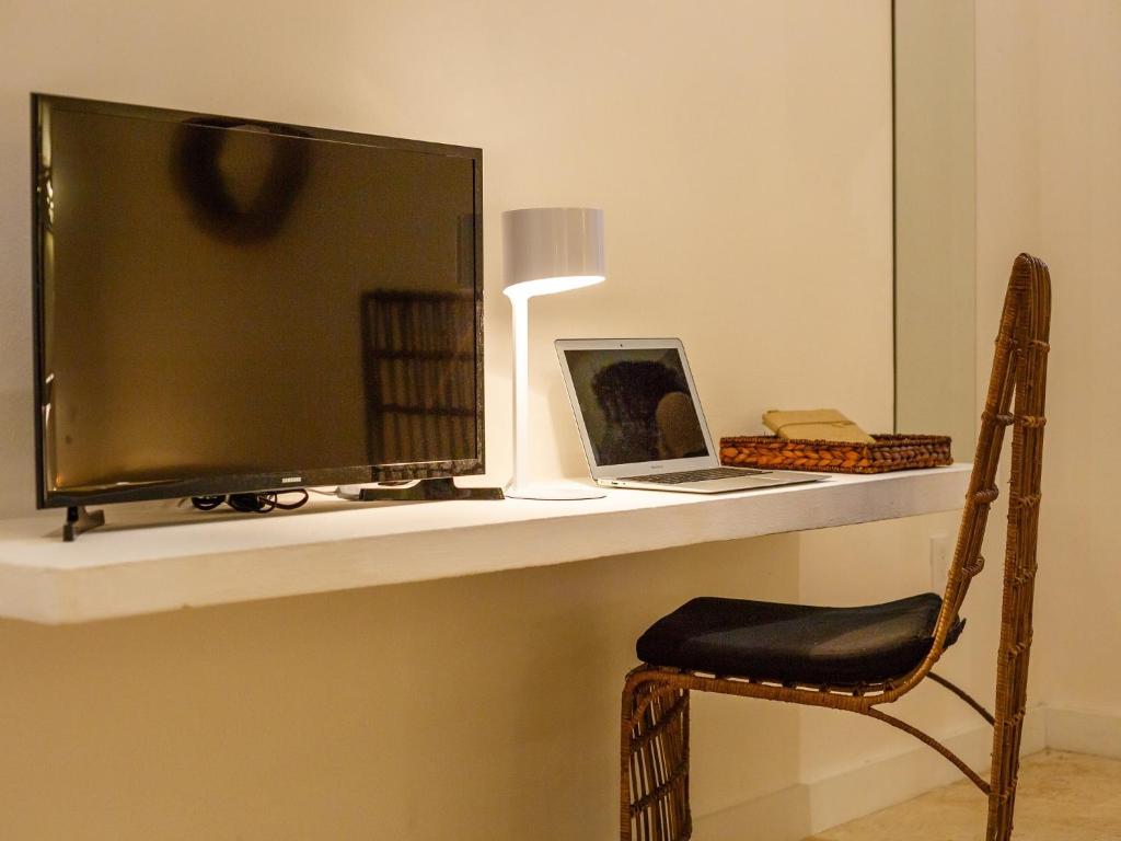 Mūsdienīga darba vieta ar televizoru, klēpjdatoru un stilīgu galda lampu uz baltas virsmas. Sēdvietā ir krēsls ar pinuma dizainu, kas piešķir telpai siltumu.