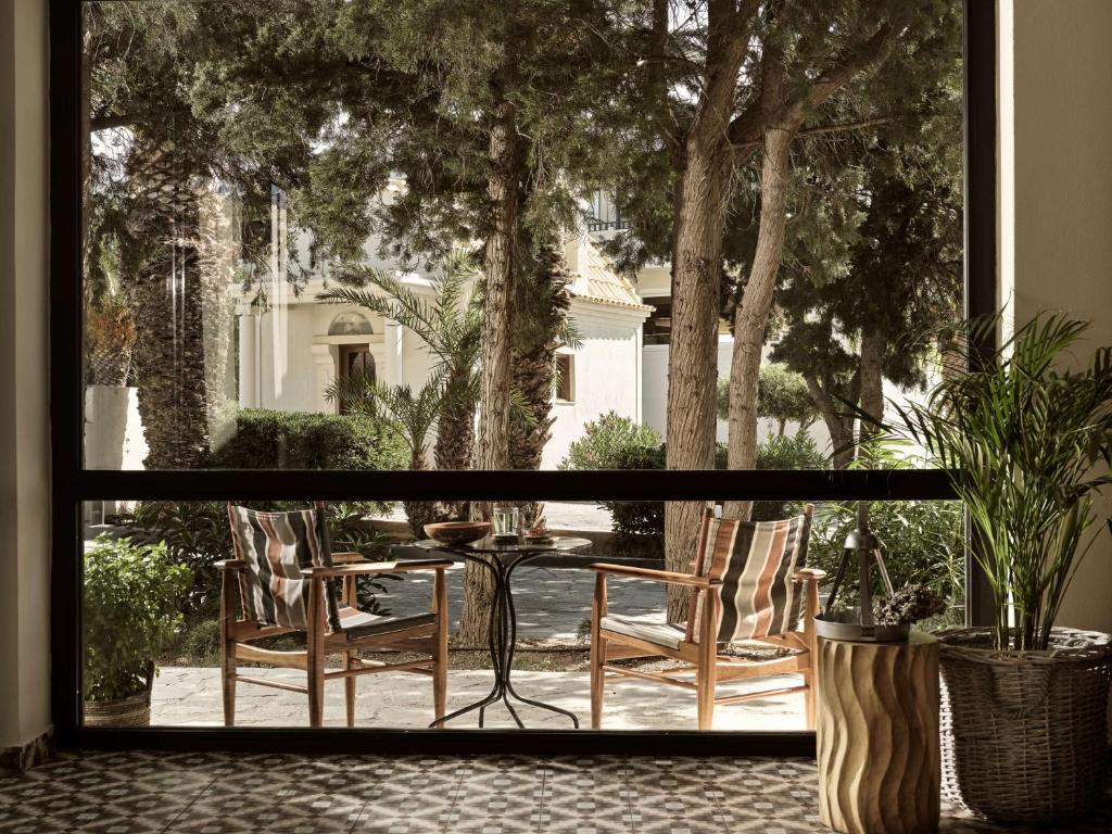 Skats no loga uz mierīgu terasi, kur redzami divi krēsli pie neliela galda, ko ieskauj zaļumi un koki. Daba un arhitektūra harmoniski saplūst.