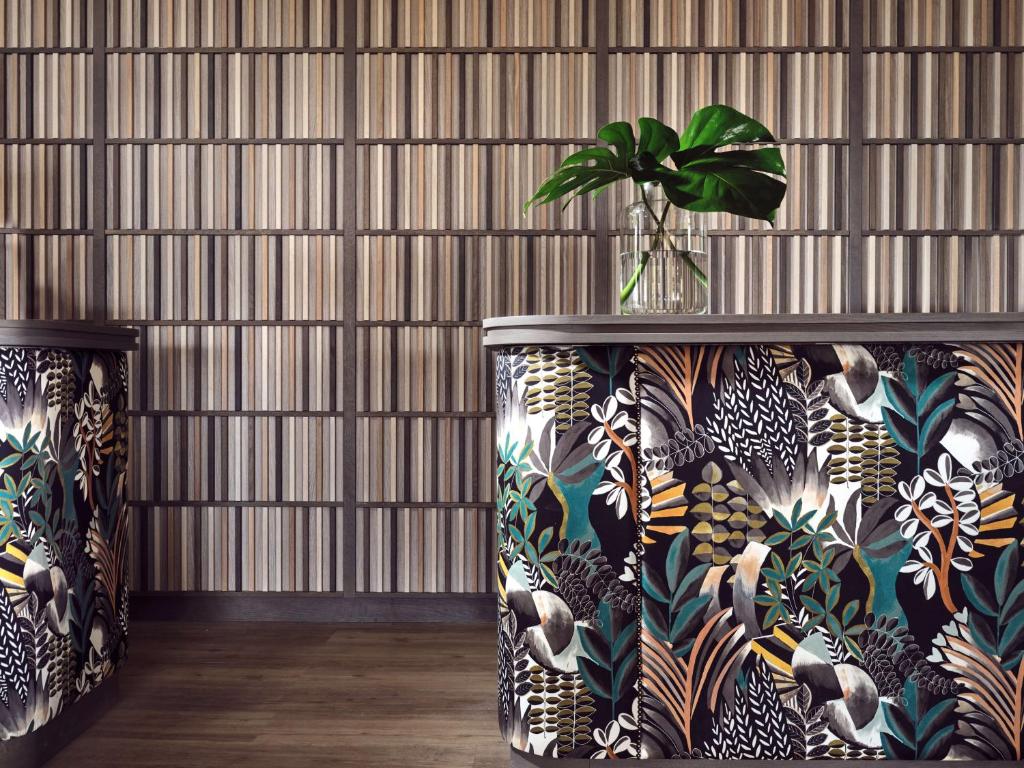 Mūsdienīgs interjers ar divām krāsainām virsmām, dekorētām ar tropiskiem rakstiem, un koka paneļu sienu fonā. Uz galda atrodas vāze ar zaļu augu.