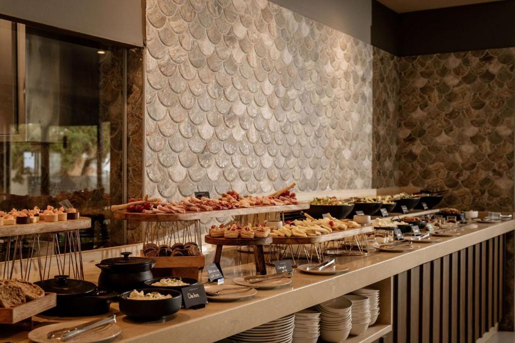 Bufete ar dažādiem ēdieniem, tostarp gaļas izstrādājumiem, salātiem un konditorejas izstrādājumiem, izvietota uz eleganta galda ar modernu dizainu. Fons ar akmens apdari piešķir siltumu un viesmīlību.