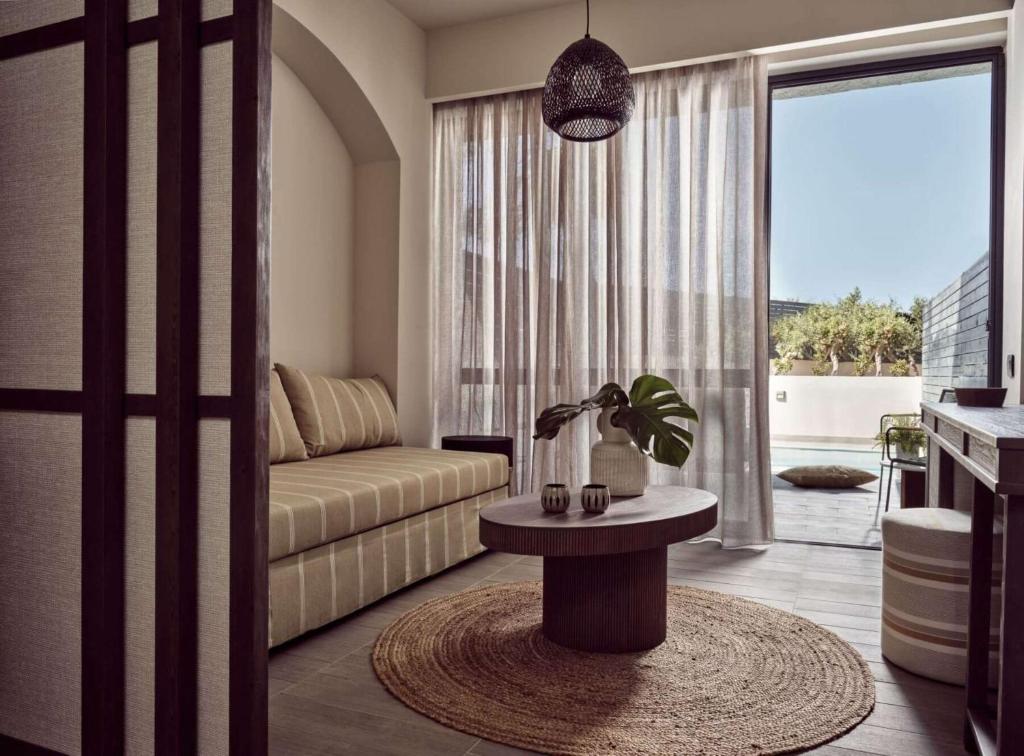 Mūsdienīgs viesistabas interjers ar dīvānu, apaļu galdu un lieliem logiem, caur kuriem redzams āra baseins un zaļumi. Eleganta un mājīga atmosfēra.