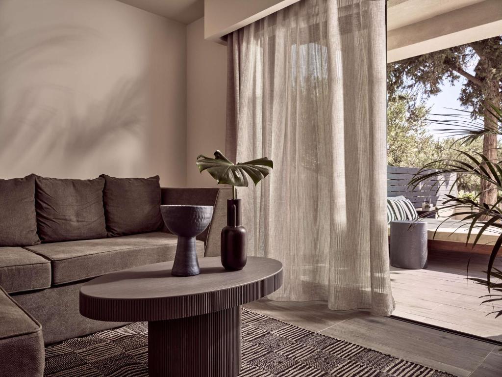 Mūsdienīgs viesistabas interjers ar dīvānu, apaļu galdu un dekoratīvām augu kompozīcijām. Gaismas caurlaidīgas aizkaru drānas rada mājīgu atmosfēru.