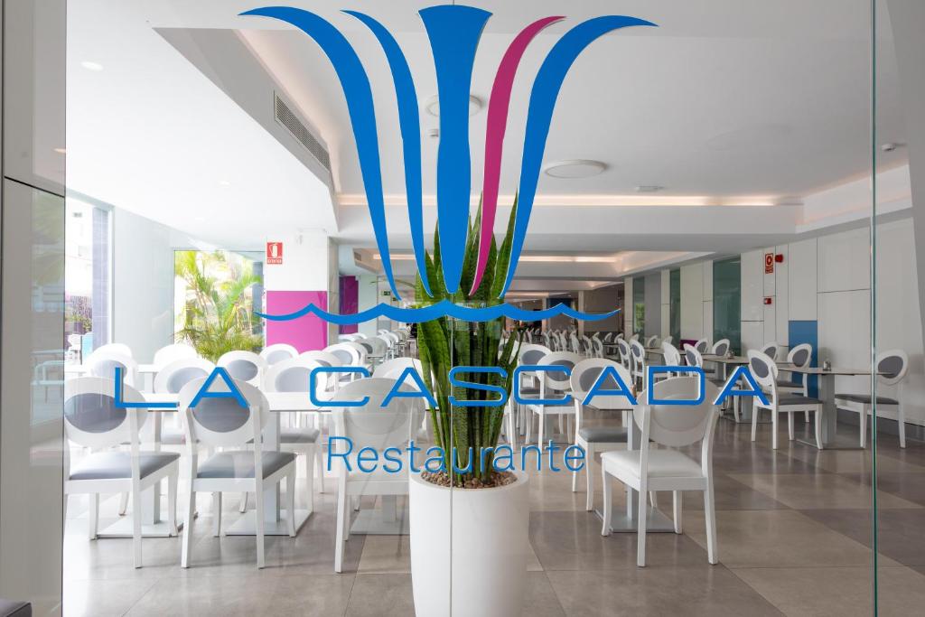 Restorāna "La Cascada" logo ar modernu interjeru, kas ietver baltas krēslus un augu, radot svaigu un gaumīgu atmosfēru.