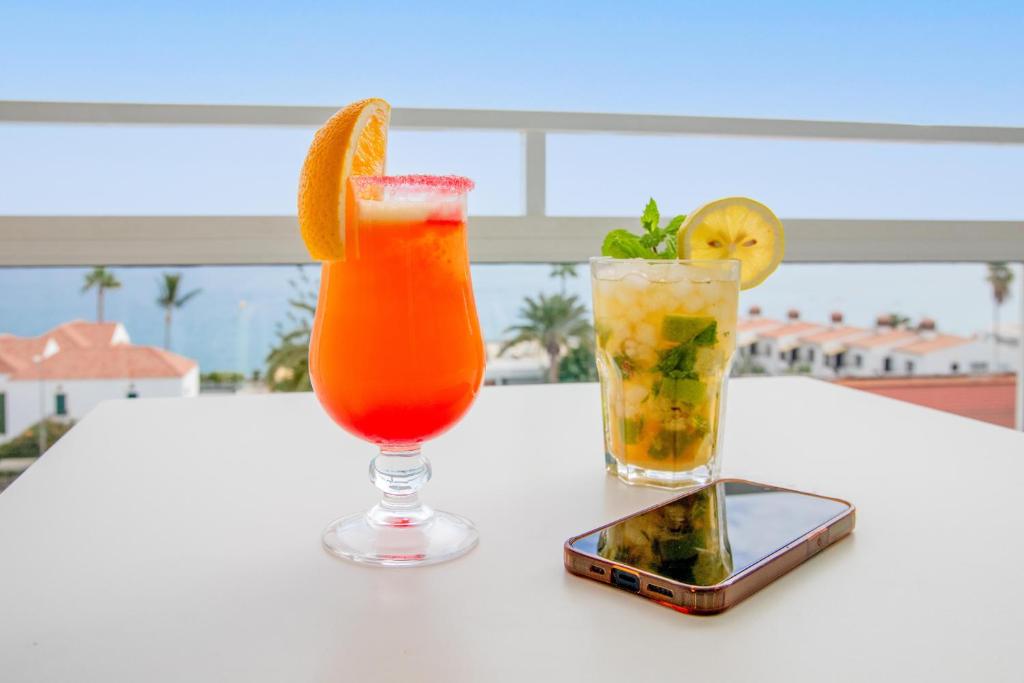 Svaigi dzērieni uz galda ar jūras skatu, iekļaujot spilgti sarkano kokteili ar apelsīnu un zaļo mojito ar citronu.