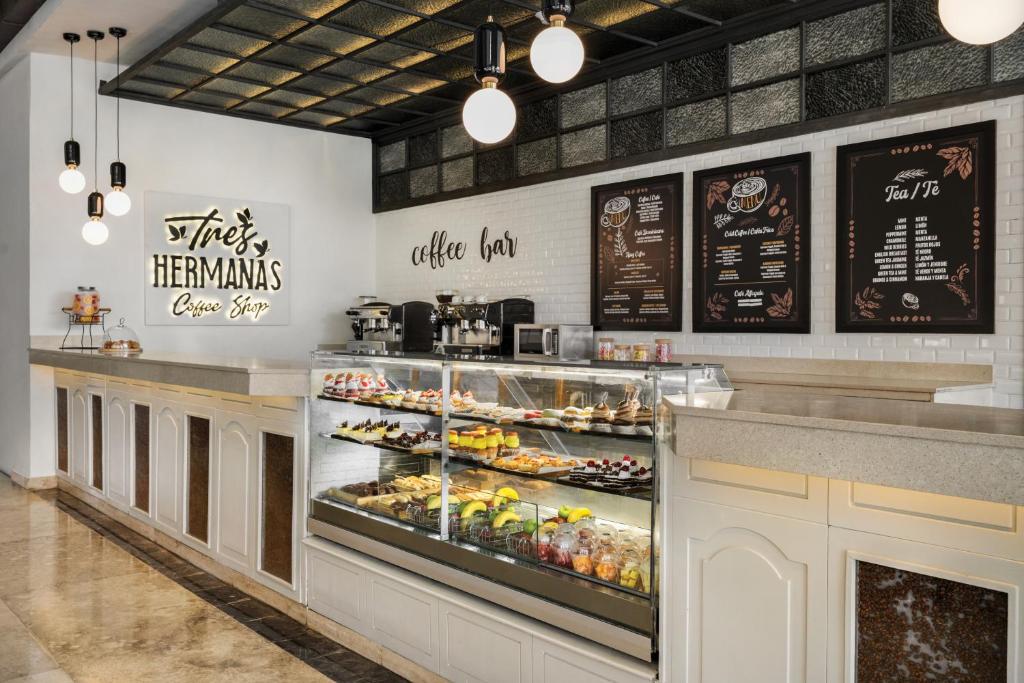 Kafijas veikala interjers ar stilīgu galdu, vitrīnu ar dažādiem konditorejas izstrādājumiem un kafijas izvēlni uz sienas. Gaismas elementi rada siltu atmosfēru.