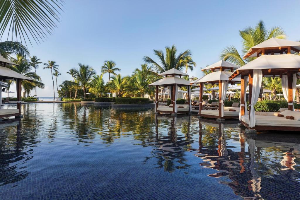 Tropu kūrorts ar palmām un atpūtas teltīm pie baseina, kas atspoguļojas ūdenī. Ideāla vieta relaksācijai un atpūtai.