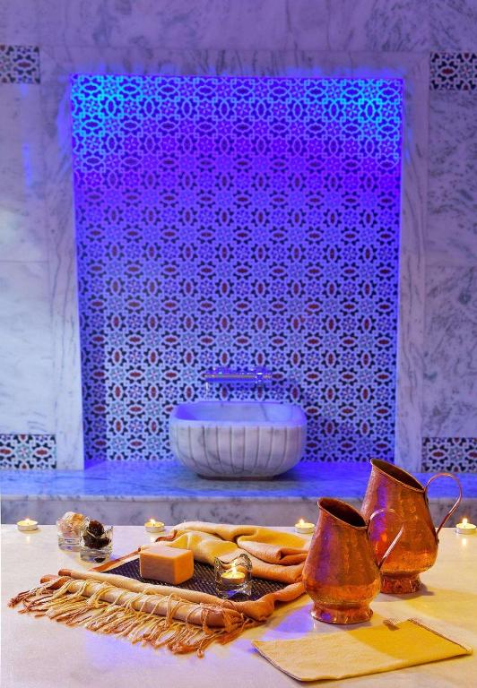 Elegants spa vidus ar dekoratīvām flīzēm un maigu apgaismojumu, kur redzami sudraba trauki un aromātiski produkti. Atmosfēra veidota relaksācijai un labsajūtai.