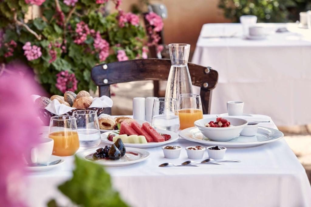 Broka veida galds ar dažādām brokastu delikatesēm, tostarp augļiem, dzērieniem un maizes izstrādājumiem, ieskauts ziedu krāšņumā.