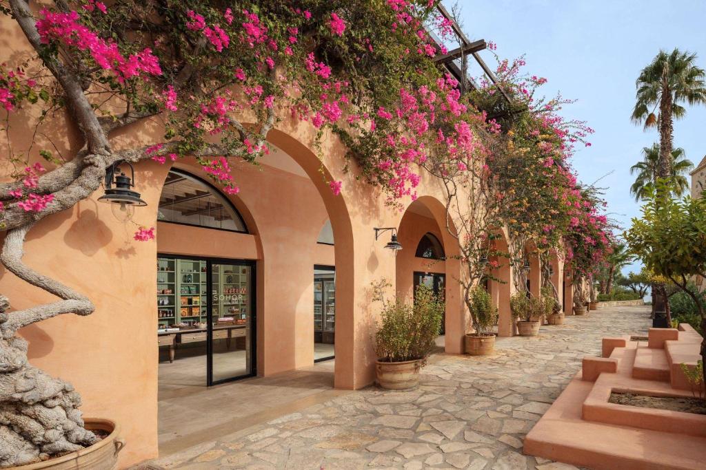 Skaista arhitektūra ar rozā ziediem, kas apvij ēku ar arku. Uz ielas redzamas akmens plāksnes un zaļas augu kompozīcijas.