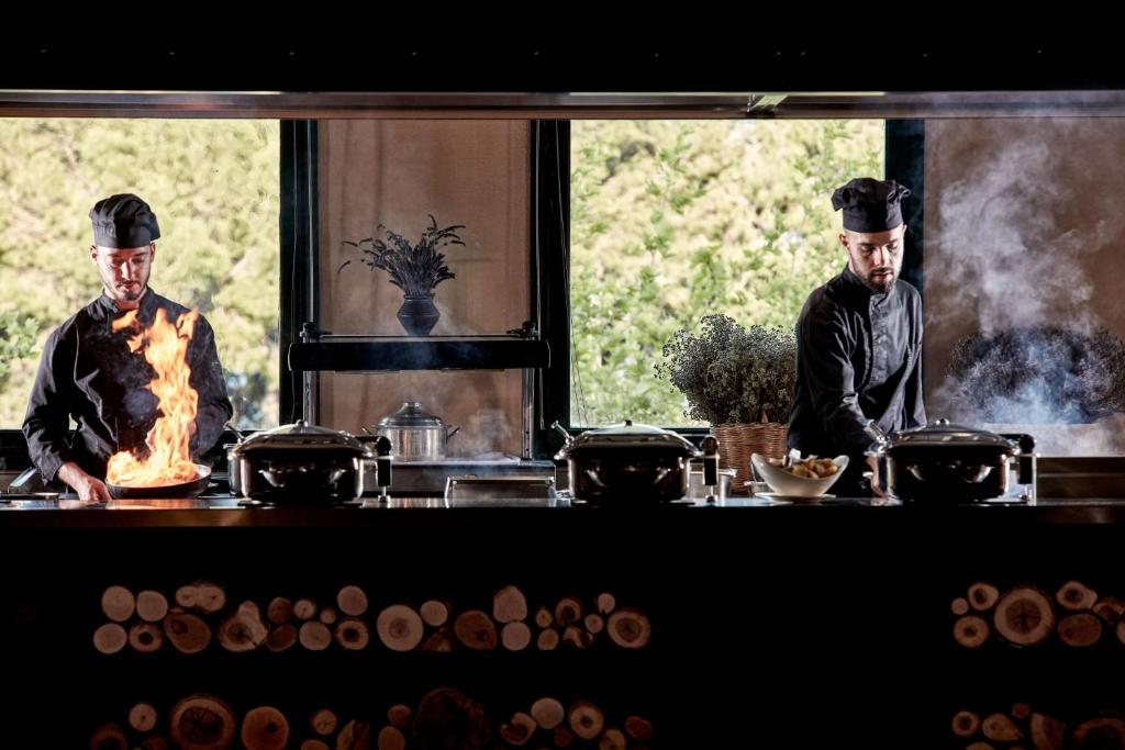 Divi šefpavāri gatavo ēdienus modernā virtuvē, kurā redzams uguns un tvaiki, radot dinamisku atmosfēru. Logi piedāvā skatu uz zaļu dabu, kas papildina kulinārijas pieredzi.