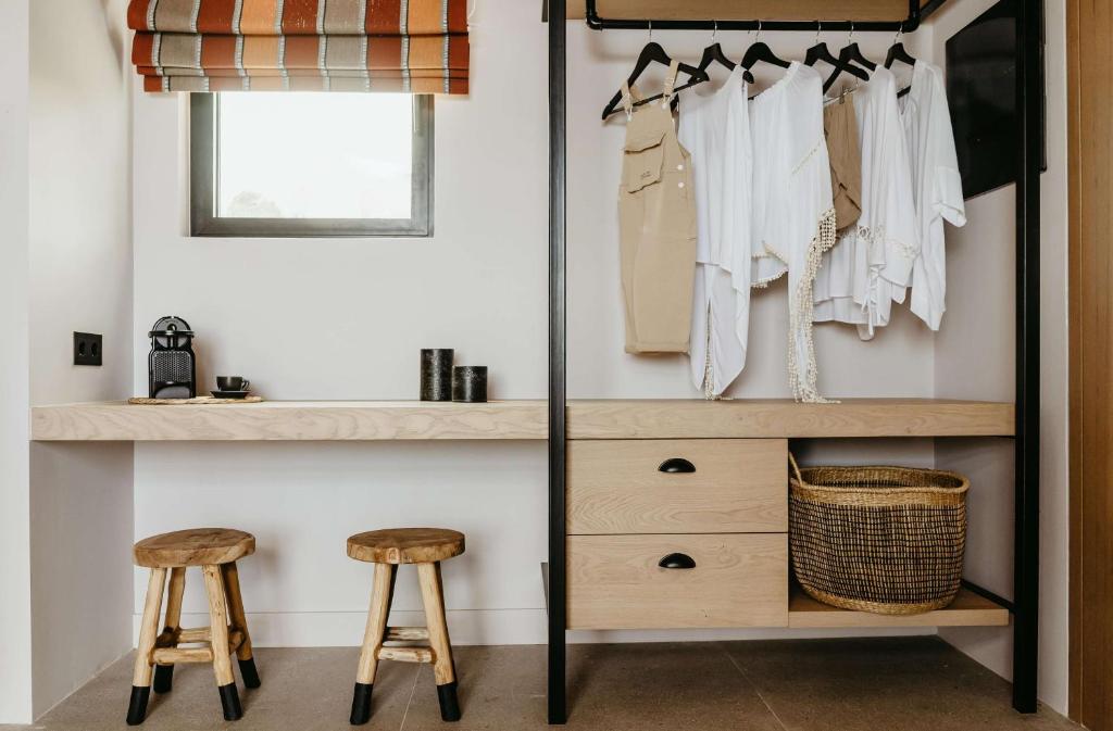 Mūsdienīgs garderobes stūris ar koka plauktu, krēslām un apģērbu pakaramajiem, kas satur baltus un smilškrāsas apģērbus. Aiz loga redzama žalūzija ar sarkanu un oranžu rakstu.